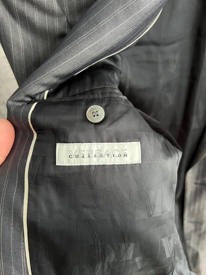 May include: A dark gray pinstriped blazer with a black interior lining featuring a repeating V logo pattern. A white rectangular label reads VERSACE COLLECTION in black text, positioned below a circular gray button with embossed branding.