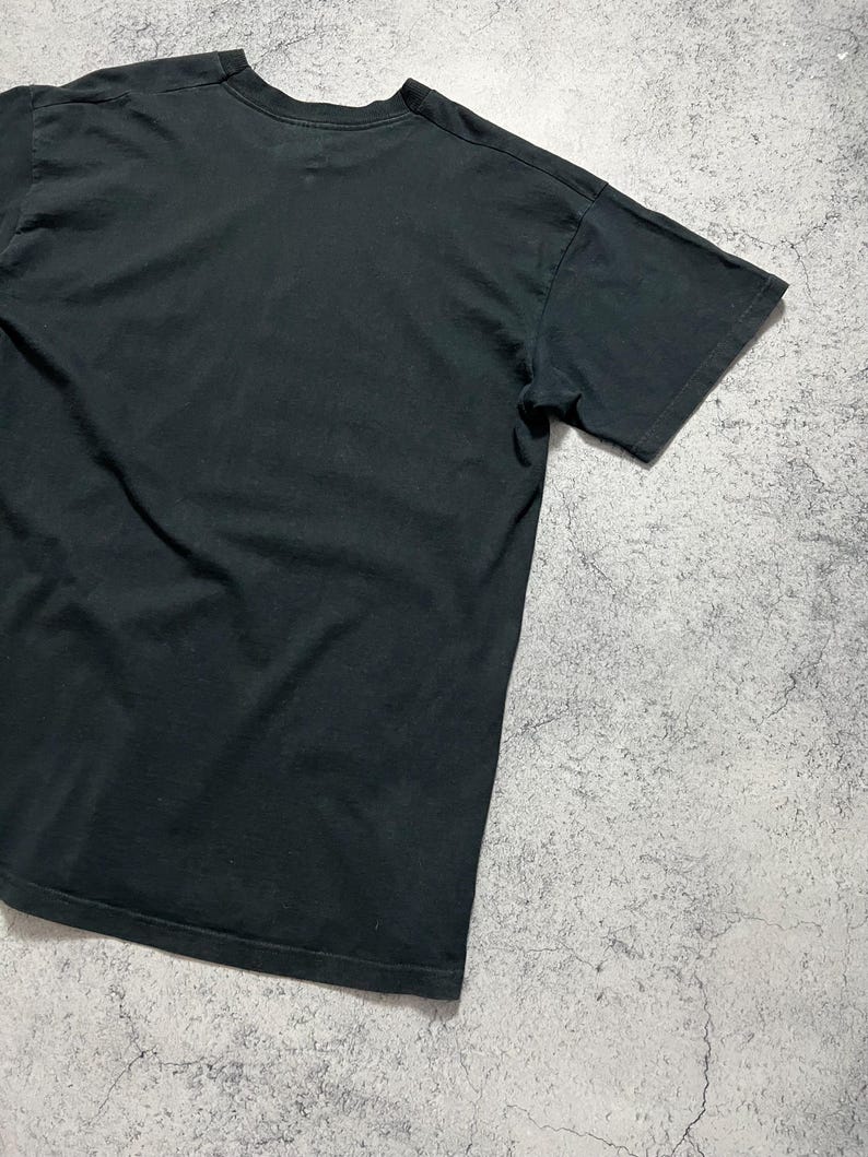 May include: A black crew neck t-shirt. The short-sleeved shirt is made of a soft material and is slightly wrinkled. The shirt is displayed on a gray and white textured surface.