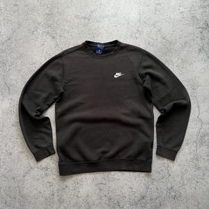 May include: A dark brown crew neck sweatshirt with the Nike logo in white on the chest. The sweatshirt has long sleeves and a ribbed hem and cuffs. The background is a mottled gray surface.