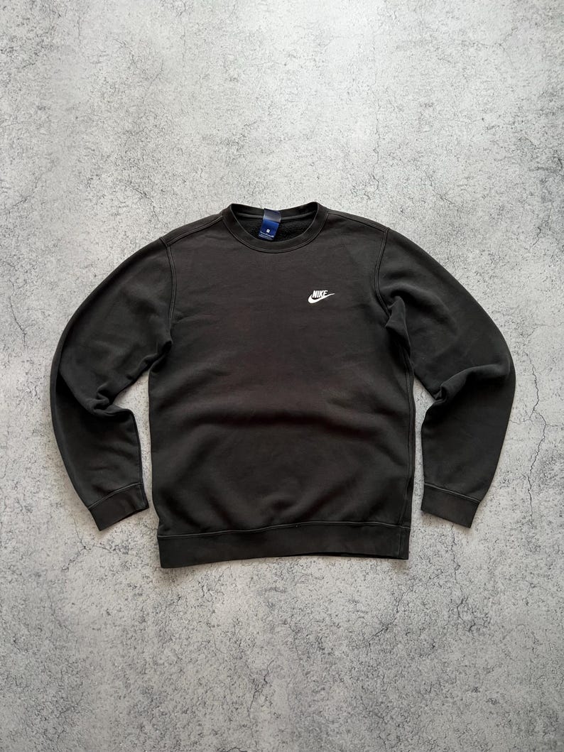 May include: A dark brown crew neck sweatshirt with the Nike logo in white on the chest. The sweatshirt has long sleeves and a ribbed hem and cuffs. The background is a mottled gray surface.