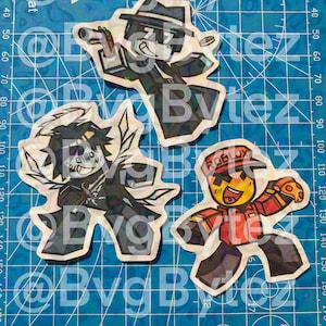 May include: Three cartoon character stickers on a blue cutting mat. The stickers feature characters in various poses, with one wearing a hat and holding a gun, another with angel wings, and a third with a Roblox hat.