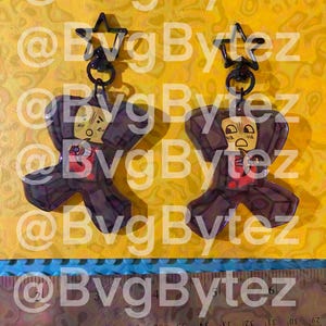May include: Two acrylic keychains featuring a cartoon character with a surprised expression, holding a small knife. Each keychain has a black star-shaped clasp. A ruler at the bottom shows the items are approximately 3 inches in total height.