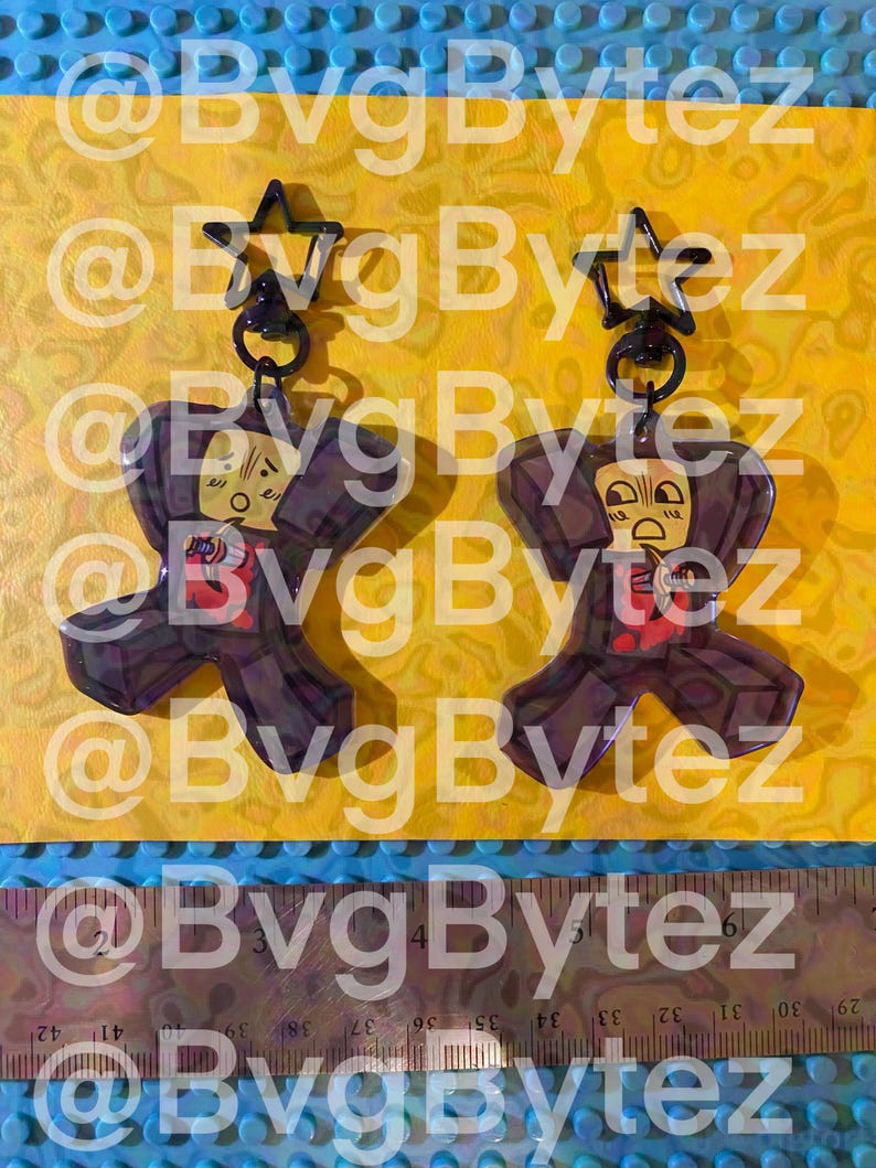 May include: Two acrylic keychains featuring a cartoon character with a surprised expression, holding a small knife. Each keychain has a black star-shaped clasp. A ruler at the bottom shows the items are approximately 3 inches in total height.