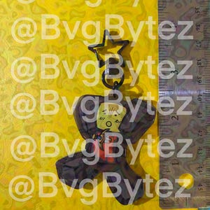 May include: A clear, dark brown acrylic keychain with a cartoon character design. The keychain has a star-shaped top and a ring. The character has a yellow face, red shirt, and a black cape. The background is yellow with the text "@BvgBytez".