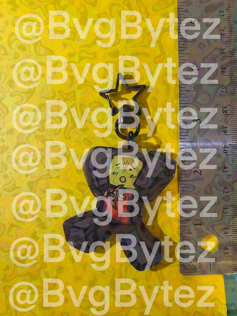 May include: A clear, dark brown acrylic keychain with a cartoon character design. The keychain has a star-shaped top and a ring. The character has a yellow face, red shirt, and a black cape. The background is yellow with the text "@BvgBytez".
