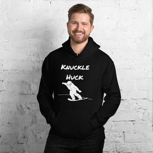 Può includere: Felpa con cappuccio nera con la scritta bianca "KNUCKLE HUCK" sopra una grafica bianca di uno snowboarder. La felpa con cappuccio ha un cappuccio con coulisse e una tasca frontale. Lo sfondo è un muro di mattoni bianchi.