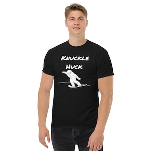 Op de afbeelding: Zwart T-shirt met de witte tekst "KNUCKLE HUCK" boven een witte afbeelding van een snowboarder. De snowboarder is in beweging, in de lucht, op een snowboard. Het T-shirt heeft korte mouwen en wordt door een persoon gedragen.