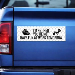 May include: A rectangular white bumper sticker with black text and graphics. The text reads "I'M RETIRED YOU'RE NOT HAVE FUN AT WORK TOMORROW." Graphics include a person relaxing in a chair and another in a beanbag chair. The background is a chain-link fence.