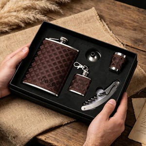 May include: A brown leather-wrapped flask set with a diamond pattern, including a small flask, shot glass, and corkscrew. The set is presented in a black box, perfect for gifting. The flask has the name "George" engraved on it.