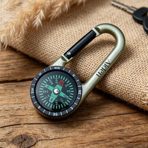 May include: A green carabiner compass with the name "Aiden" printed on the side. The compass has a black and green face with directional markings. A set of keys rests on a burlap cloth in the background.