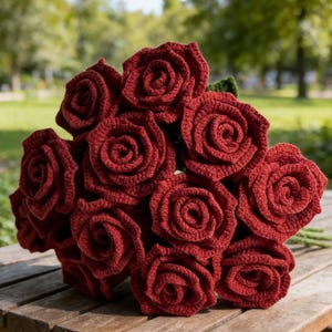 May include: A bouquet of handmade, crocheted roses in a deep red colour. The roses are arranged in a cluster, with green stems and leaves visible. The bouquet is placed on a wooden surface, with a blurred background of trees.