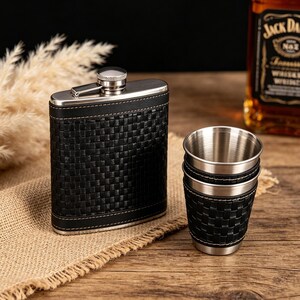 Peut inclure: Une flasque en cuir noir avec un bouchon en acier inoxydable et un ensemble de trois verres à shot en acier inoxydable avec des revêtements en cuir noir assortis. Sur une surface en bois.