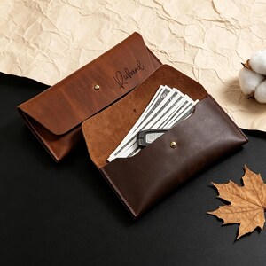 May include: Two brown leather wallets, one open to reveal cash and a car key. The other wallet is closed with the name "Richard" inscribed. Both wallets have a snap closure. A cotton flower and a leaf are also in the image.
