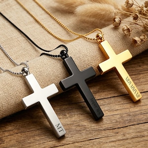 May include: Three cross pendant necklaces in silver, black, and gold. Each cross is engraved with text, including initials and a date in Roman numerals. The necklaces rest on a tan fabric and wooden surface.
