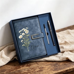 Op de afbeelding: Een blauw leren notitieboek met een bloemenontwerp en de naam "Oliver" in script, gepresenteerd in een bijpassende blauwe geschenkdoos met twee pennen. Het notitieboek heeft een goudkleurige sluiting.