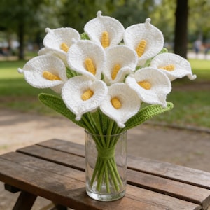 Könnte beinhalten: Ein Strauß gehäkelter Calla-Lilien in einer durchsichtigen Glasvase. Die Blumen sind weiß mit gelben Zentren, grünen Stielen und grünen Blättern. Die Vase steht auf einem Holztisch.