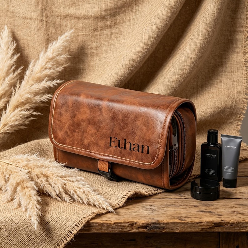 May include: A brown leather-look toiletry bag with a fold-over flap and a buckle closure. The name Ethan is embossed in black on the front. Beside the bag are a black bottle, a black tube, and a small black jar on a wooden surface.