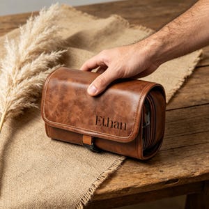 May include: A brown leather toiletry bag with a smooth, textured finish and a flap closure. The name Ethan is embossed in black lettering on the front flap. The bag features a small brown strap with a black buckle detail on the lower front section.