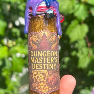 May include: A small, glittery, gold-colored bottle with a purple and blue top. The label reads "DUNGEON MASTER'S DESTINY" with dragon and treasure chest graphics. A black gem and red string are on the top. The bottle is held in a hand.