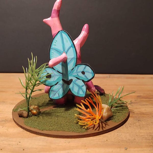 May include: A miniature diorama featuring a stylized coral reef scene. A pink coral structure supports a large, blue-green leaf-like form. The base includes green grass, small rocks, and orange coral, creating a vibrant underwater aesthetic.
