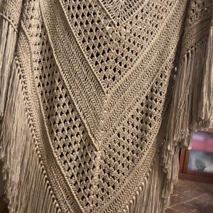 Può includere: Uno scialle triangolare all'uncinetto color beige caratterizzato da un intricato motivo traforato con punti pieni e a forma di diamante alternati. Il capo è rifinito con lunghe frange dritte lungo i bordi, creando un aspetto strutturato.
