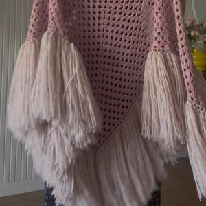 Op de afbeelding: Een driehoekige gehaakte sjaal in een oudroze tint, voorzien van een opengewerkt granny square patroon over het hele lichaam. De randen zijn versierd met lange, dikke franjes van lichtroze garen die verticaal langs de onderkant hangen.