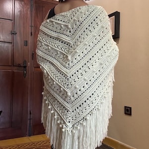 Pode incluir: Xale de crochê feito à mão, de cor creme e formato triangular. O xale apresenta intrincados padrões vazados, bolinhas texturizadas e uma franja de longas borlas na borda inferior. Um acessório aconchegante e estiloso.