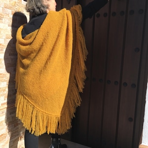Puede incluir: Poncho amarillo mostaza con flecos. El poncho está hecho de una tela de punto gruesa y texturizada. Los flecos cuelgan a lo largo del borde inferior. La persona lleva pantalones negros y botas negras. El fondo incluye una puerta de madera y una pared de ladrillo.