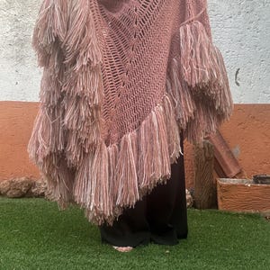 Peut inclure: Un châle en crochet rose poudré présentant un motif en V ajouré et de longues franges épaisses sur les bords. Le fil est un mélange de fibres rose sourd et blanc, créant un aspect texturé sur l'ensemble du vêtement triangulaire.