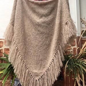 Pode incluir: Um poncho bege de malha com detalhes de franjas. O poncho é levantado, mostrando sua forma triangular e malha texturizada. As franjas pendem ao longo da borda inferior e das mangas, adicionando um elemento decorativo.