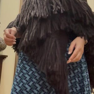 Peut inclure: Un châle à franges marron foncé. Le châle a un col haut et plusieurs couches de franges. Le vêtement est fait d'une matière douce et texturée. Le châle est porté sur une jupe à motifs bleus et verts.