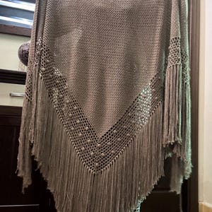 Peut inclure: Un châle triangulaire de couleur taupe présentant une section centrale en tricot dense, une bordure ajourée décorative avec de petits détails en relief et de longues franges droites le long des bords inférieurs. Le vêtement est tenu en hauteur.