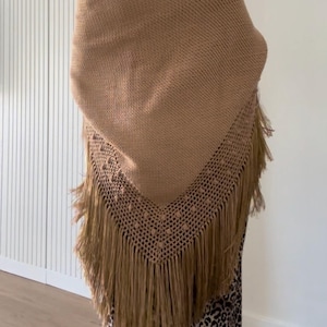 Puede incluir: Un chal triangular de color tostado con una sección superior de punto denso y un borde calado decorativo con pequeñas borlas en relieve. El borde inferior está rematado con largos flecos rectos que cuelgan de la base puntiaguda.