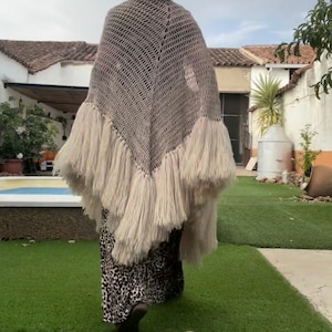 Può includere: Uno scialle all'uncinetto beige con un motivo triangolare a maglia aperta e lunghe frange spesse lungo il bordo inferiore. Lo scialle è indossato sopra una gonna lunga con stampa leopardata bianca e nera che arriva fino alle gambe.