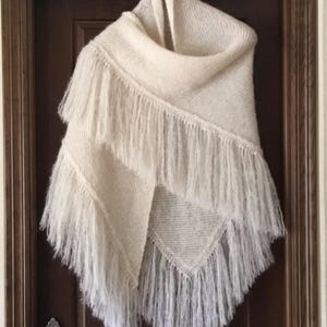 Può includere: Sciarpa lavorata a maglia color crema con lunghe frange. La sciarpa è appesa a un gancio su una porta marrone. La sciarpa è realizzata con un filato morbido e strutturato e ha una forma triangolare.