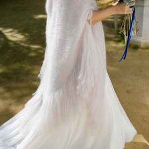 Pode incluir: Um vestido de noiva branco com uma saia fluida e um xale macio com franjas. O xale é drapeado sobre os ombros e os braços. Um buquê de vegetação e flores brancas é segurado na mão.