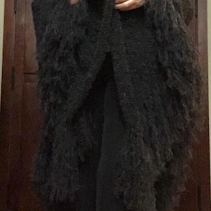 Könnte beinhalten: Ein langer, dunkelgrauer Schal oder Wickel mit Fransen. Das Kleidungsstück hat ein strukturiertes, zotteliges Aussehen und wird über einem helleren Oberteil getragen. Der Schal fällt über die Vorderseite und erzeugt einen geschichteten Effekt. Der Gesamtstil ist elegant und gemütlich.