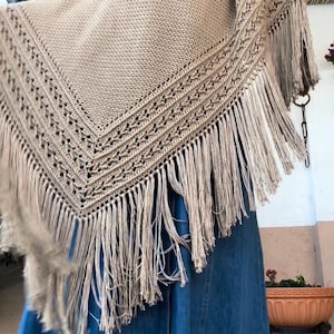 Puede incluir: Un chal de ganchillo beige con un borde con flecos. El chal tiene forma triangular con un diseño de borde detallado. La franja cuelga, agregando textura y movimiento a la prenda. El chal está drapeado sobre una prenda de mezclilla azul.