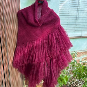 Pode incluir: Um xale de tricô em tom magenta profundo com gola alta e um design de franjas texturizadas em camadas que cai diagonalmente pela frente. A peça é confeccionada com fio macio e felpudo e está pendurada em um cabide contra uma parede.