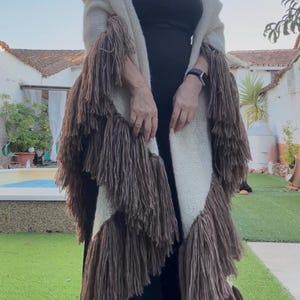 Puede incluir: Un chal de punto color crema con largos flecos marrones en capas a lo largo de los bordes. La prenda se lleva sobre una camiseta negra sin mangas, mostrando una construcción de punto texturizado y borlas de hilo marrón en cascada al frente.