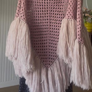 Può includere: Uno scialle triangolare all'uncinetto in una tonalità rosa polvere con un motivo a rombi traforato. I bordi dello scialle sono decorati con lunghe e spesse frange di filato rosa chiaro che pendono dai lati e dalla punta inferiore del capo.