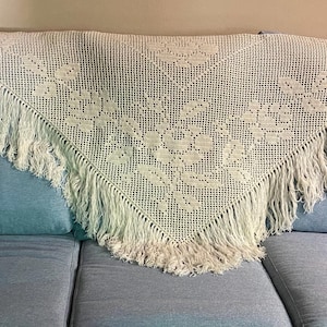 Peut inclure: Châle triangulaire en crochet crème avec des motifs floraux et des franges. Le châle est drapé sur un canapé bleu clair avec des coussins assortis. Le design et la texture complexes sont visibles.