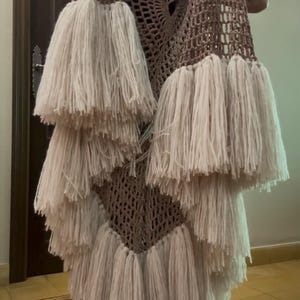 Op de afbeelding: Een mouwloos, beige gehaakt kledingstuk met een open, netachtig patroon over het hele lichaam. Het kledingstuk is versierd met meerdere lagen lange, dikke, crèmekleurige garenfranjes die aan de onderkant van de stof naar beneden hangen.