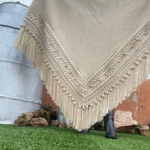 Peut inclure: Un châle triangulaire beige avec des détails de crochet complexes et une bordure à franges. Le châle est présenté, mettant en valeur sa texture et son design. L'arrière-plan comprend un récipient en métal, des rochers et de l'herbe verte.