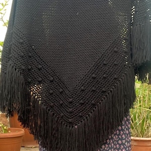 Puede incluir: Un poncho negro de crochet con dobladillo con flecos. El poncho presenta un diseño de cuello en V con bolitas texturizadas y flecos largos. Este accesorio hecho a mano es perfecto para añadir un toque bohemio a cualquier atuendo.