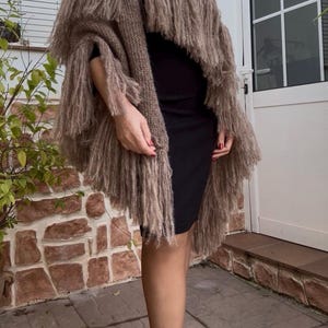 Peut inclure: Un poncho tricoté de couleur taupe présentant plusieurs couches de longues franges ébouriffées. Le vêtement est drapé sur une robe noire simple, mettant en valeur une construction en fil texturé et duveteux avec un ourlet asymétrique.
