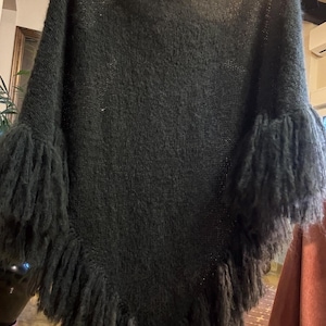 Puede incluir: Poncho de punto verde oscuro con flecos. El poncho tiene un diseño holgado y bordes con flecos en el dobladillo y las mangas. El tejido de punto parece suave y texturizado, proporcionando calidez y estilo. Una prenda cómoda y de moda.