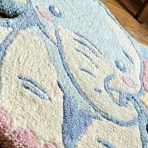 May include: A whimsical, tufted rug in the shape of a cartoon bunny. The rug features a light blue, cream, pink, and yellow color palette. The bunny's face and ears are clearly defined, creating a playful and inviting design.