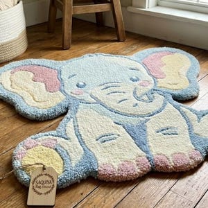 May include: A light blue, white, pink, and yellow elephant-shaped rug. The rug has a tufted design with a smiling face and large ears. The rug is on a hardwood floor. A tag is attached to the rug.