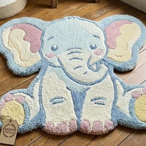 May include: A handmade, tufted rug in the shape of a friendly elephant. The rug features a light blue body, pink and yellow accents, and a cream-colored face and legs. The rug is placed on a wooden floor.